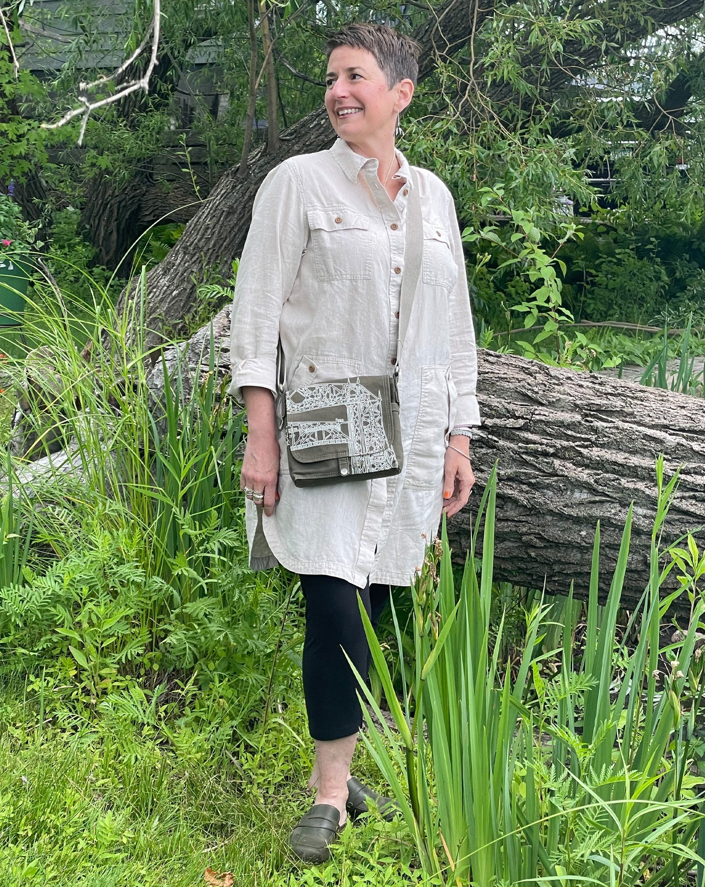 Dock 5’s hand-crafted Aerial Lift Bridge Canvas Messenger Bag in olive is being worn by a smiling woman standing near the shoreline of Park Point