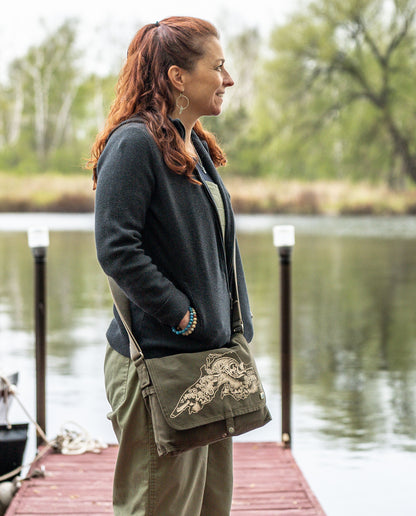Dock 5’s hand-crafted Lake Superior Canvas Messenger Bag in olive is being worn by a smiling woman near a lake