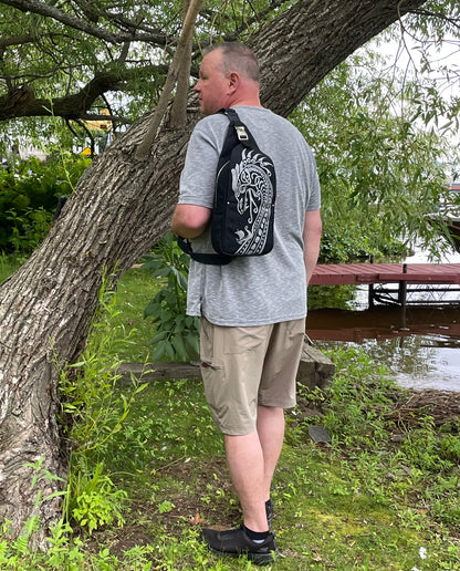 Dock 5’s hand-crafted Viking Dragon Canvas Sling Bag in black is being worn by a man standing near the shoreline of Park Point